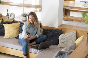 Mid adult woman using smart phone while sitting with laptop on couch in living room - SBOF03724