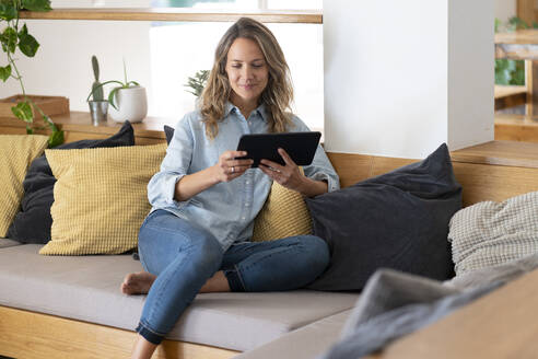 Entspannte Frau, die ein Video über ein Tablet ansieht, während sie auf der Couch im Wohnzimmer sitzt - SBOF03725