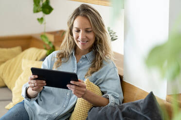Lächelnde Frau, die ein digitales Tablet benutzt, während sie zu Hause auf der Couch im Wohnzimmer sitzt - SBOF03726