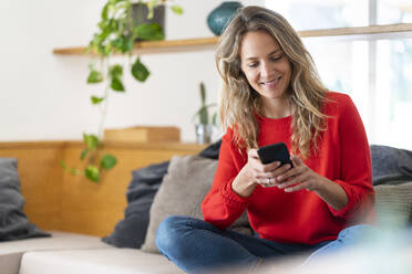 Lächelnde blonde Frau Textnachrichten auf dem Handy, während sie im Wohnzimmer zu Hause sitzt - SBOF03728