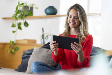 Lächelnde blonde Frau, die ein Tablet benutzt, während sie auf der Couch im Wohnzimmer sitzt - SBOF03729