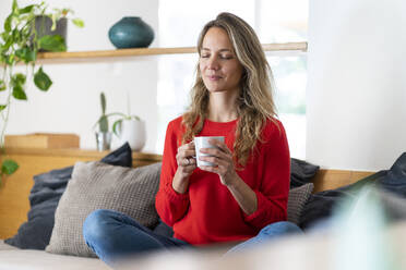 Mid adult woman holding coffee cup while sitting with eyes closed in living room at home - SBOF03730