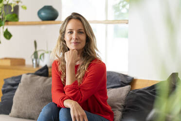 Confident blond woman sitting with hand on chin in living room at home - SBOF03731