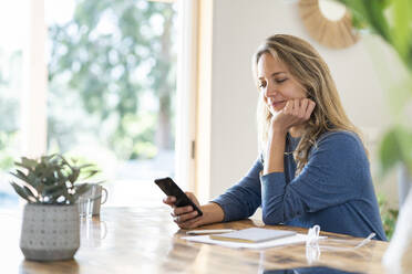 Blonde Unternehmerin, die am Tisch sitzend eine Textnachricht über ihr Mobiltelefon verschickt - SBOF03736