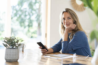 Smiling female professional sitting with mobile phone at table while looking away - SBOF03737