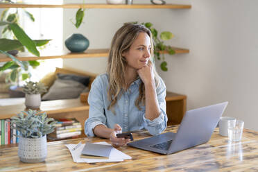 Female professional sitting with mobile phone and laptop at table while looking away - SBOF03745