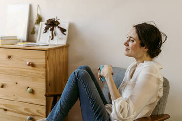 Smiling woman with cup looking away while sitting on chair at home - VABF04293