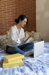 Frau, die ein Mobiltelefon benutzt, während sie zu Hause im Schlafzimmer am Laptop sitzt - VABF04302