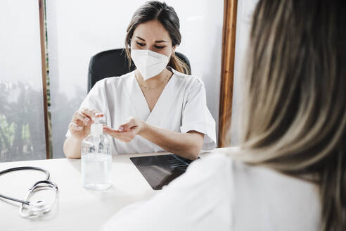 Arzt mit Gesichtsschutzmaske nimmt Desinfektionsmittel am Schreibtisch in einer Klinik ein - EBBF03225