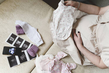 Schwangere Frau mit Ultraschalluntersuchung hält Babytücher, während sie zu Hause sitzt - EYAF01568