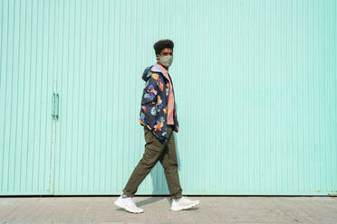 Man with hands in pockets wearing protective face mask while walking by turquoise wall - AFVF08542