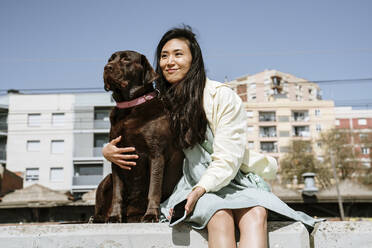 Lächelnde Frau sitzt mit Chocolate Labrador auf einer Stützmauer vor einem Gebäude - AFVF08551