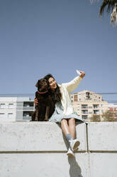 Lächelnde Frau, die ein Selfie mit ihrem Labrador-Hund macht, während sie auf einer Stützmauer sitzt - AFVF08552
