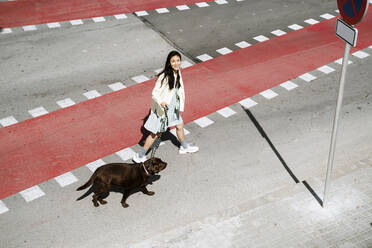 Smiling woman crossing street with Labrador dog on street - AFVF08557