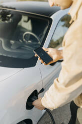 Man using charging app on smart phone while charging electric car - GMCF00132