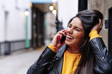 Cheerful young woman squinting while talking on mobile phone in city - ASGF00203