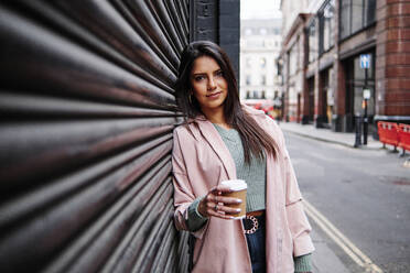 Smiling woman with coffee cup leaning on shutter in city - ASGF00207