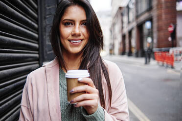 Smiling young woman with disposable coffee cup standing by shutter in city - ASGF00209
