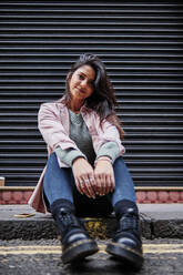 Lächelnde Frau, die vor einem Fensterladen auf dem Fußweg sitzt - ASGF00214