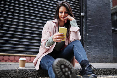 Beautiful woman with coffee cup using mobile phone while sitting on sidewalk - ASGF00216