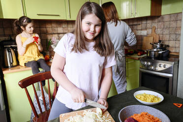 Lächelndes Mädchen, das Kohl schneidet, während Mutter und Schwester im Hintergrund Essen zubereiten - EYAF01582