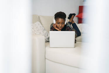 Young businesswoman with headphones working on laptop while lying on sofa at home - MPPF01616