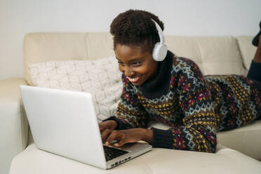 Smiling female entrepreneur with headphones working on laptop in living room - MPPF01617