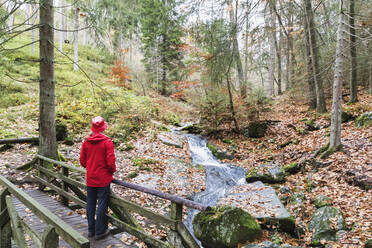 Ältere Frau mit rotem Mantel und Hut steht auf einer Brücke im Wald - GWF06969