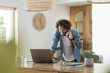 Männlicher Unternehmer, der einen Laptop benutzt, während er zu Hause telefoniert - SBOF03755