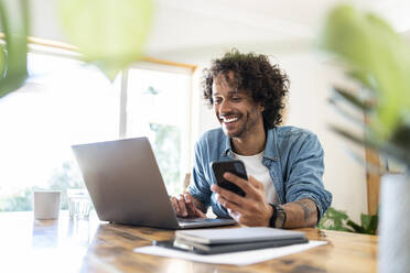 Lächelnder männlicher Berufstätiger, der ein Smartphone hält, während er einen Laptop auf einem Tisch zu Hause benutzt - SBOF03756