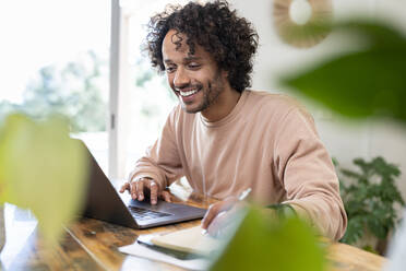 Cheerful businessman writing in diary while using laptop at home - SBOF03768