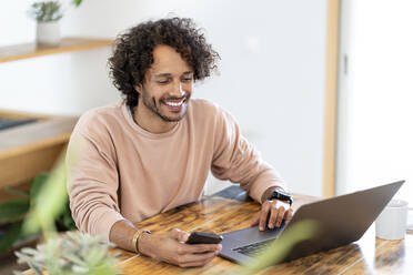 Lächelnder Geschäftsmann, der sein Smartphone hält, während er zu Hause am Laptop arbeitet - SBOF03774