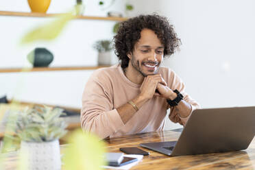 Smiling man with hands clasped looking at laptop on table at home - SBOF03775