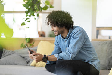 Lächelnder männlicher Fachmann, der ein digitales Tablet benutzt, während er zu Hause auf dem Sofa im Wohnzimmer sitzt - SBOF03786