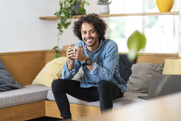Mann hält Kaffeetasse, während er zu Hause auf dem Sofa sitzt - SBOF03788