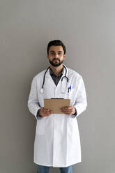 Männlicher Arzt mit Klemmbrett vor einer Wand stehend - GIOF12203