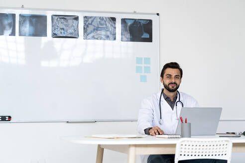 Lächelnder männlicher Arzt sitzt mit Laptop auf dem Schreibtisch im Krankenhaus - GIOF12211