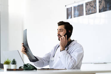 Männlicher Angestellter im Gesundheitswesen, der Röntgenbilder untersucht, während er im Krankenhaus mit seinem Smartphone telefoniert - GIOF12214