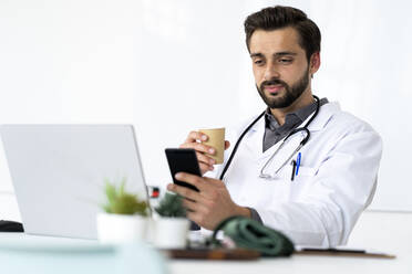 Handsome male healthcare worker drinking coffee while using mobile phone - GIOF12216