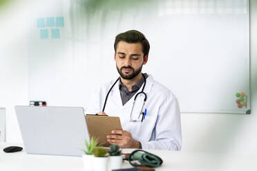 Male healthcare worker with clipboard and laptop sitting at desk - GIOF12220