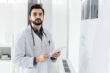 Männlicher medizinischer Experte, der ein digitales Tablet hält, während er an einer weißen Tafel im Krankenhaus steht - GIOF12243