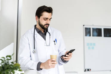 Male doctor holding disposable coffee cup while using smart phone in hospital - GIOF12249