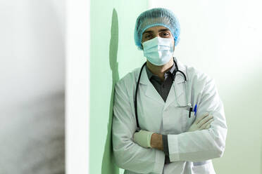 Male doctor wearing surgical cap standing with arms crossed at hospital during COVID-19 - GIOF12257