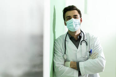 Male healthcare worker wearing protective face mask standing with arms crossed leaning on wall - GIOF12258