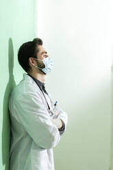 Männlicher medizinischer Experte mit Gesichtsmaske steht mit den Armen an der Wand - GIOF12259