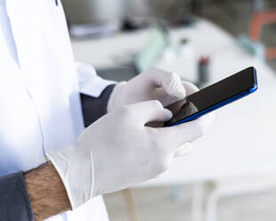 Männlicher medizinischer Angestellter mit chirurgischen Handschuhen, der im Krankenhaus eine Textnachricht über sein Mobiltelefon sendet - GIOF12270