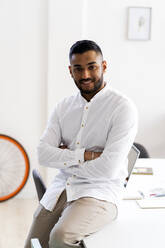 Businessman with arms crossed sitting on desk at office - GIOF12272