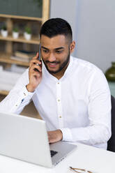 Männlicher Unternehmer, der mit seinem Smartphone telefoniert, während er einen Laptop im Büro benutzt - GIOF12278