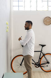 Junger Geschäftsmann schaut auf Haftnotizen an der Wand im Büro - GIOF12283