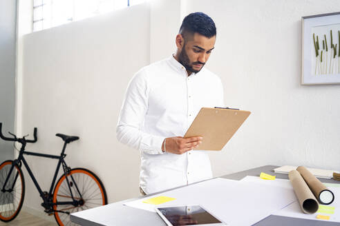 Männlicher Unternehmer, der ein Klemmbrett hält, während er in der Nähe des Schreibtischs an der Wand im Büro steht - GIOF12298